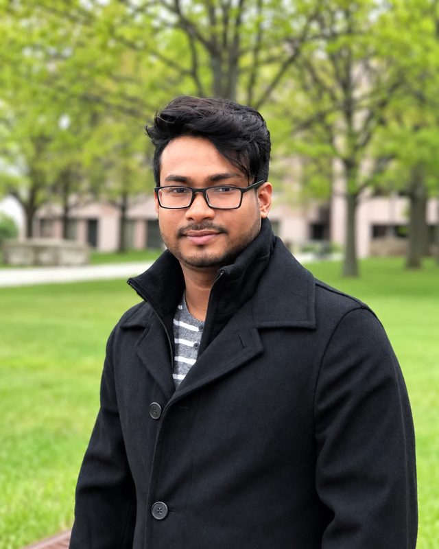 Portrait of Shekh Solayman outdoors, wearing glasses and a dark coat, standing on a tree-lined lawn